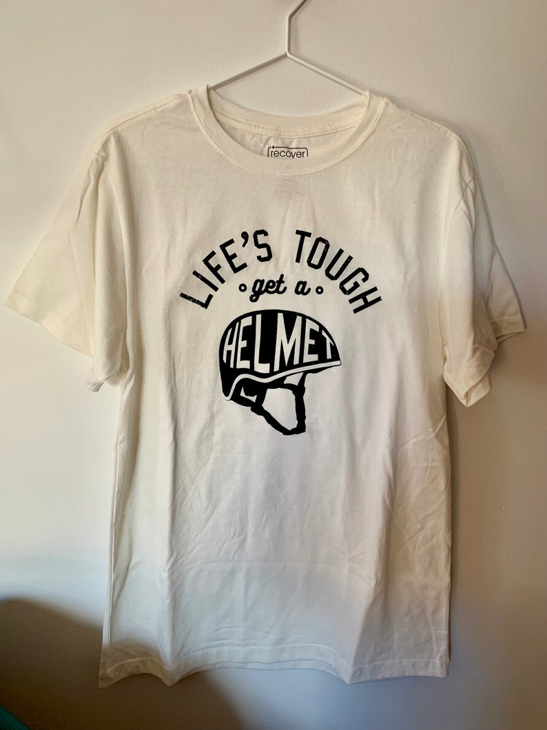 Op de afbeelding: Wit T-shirt met zwarte tekst die "Life's Tough get a HELMET" zegt met een afbeelding van een helm.