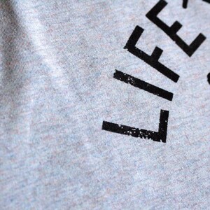 Op de afbeelding: Grijs T-shirt met zwarte tekst die "Life's a journey, get lost." luidt.