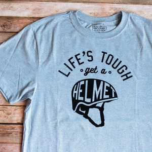 Op de afbeelding: Een lichtgrijs T-shirt met een zwarte afbeelding waarop staat "Life's Tough... get a Helmet" met een zwart silhouet van een helm.