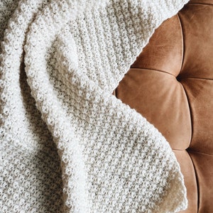 May include: A white crocheted throw blanket with a textured pattern, draped over a brown leather couch.