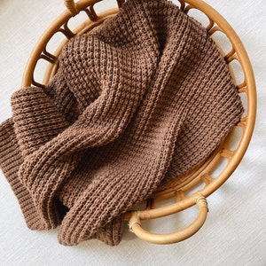 Könnte beinhalten: Eine braune Strickdecke in einem runden Weidenkorb mit zwei Griffen.