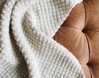 May include: A white crocheted throw blanket with a textured pattern, draped over a brown leather couch.