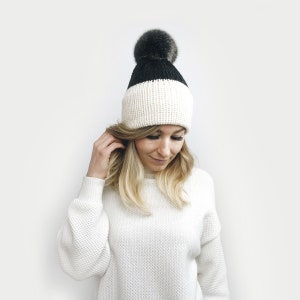 May include: A white and black knit beanie with a black faux fur pom pom. The beanie is folded down to show a white knit band.