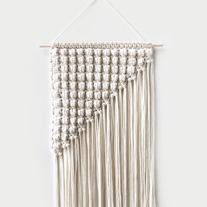 Puede incluir: Un tapiz de macramé blanco con un patrón geométrico y flecos largos. El tapiz está sujeto a una varilla de madera.