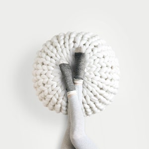 May include: A white, chunky knit pouf with grey socks and light grey leggings. The pouf is round and has a textured surface. The socks have grey and white details. The legs are crossed.