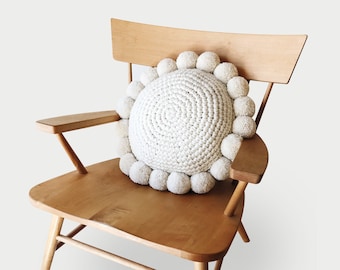 Puede incluir: Un cojín redondo de crochet blanco con ribete de pompones colocado en una silla de madera.