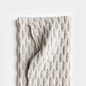 Peut inclure: Une couverture en tricot de couleur crème avec un motif en losange. La couverture est pliée en deux et les bords sont visibles.