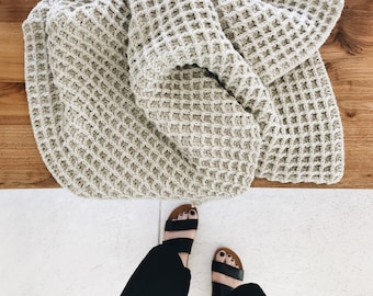 May include: A beige waffle knit throw blanket folded on a wooden surface. The blanket has a textured waffle stitch pattern.