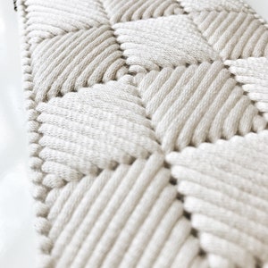 May include: A close-up of a white woven wall hanging with a diamond pattern. The hanging is made of thick, textured rope and has a simple, modern design.