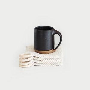 May include: A black ceramic mug with a handle sits on top of a stack of four white crocheted coasters with wooden rings.