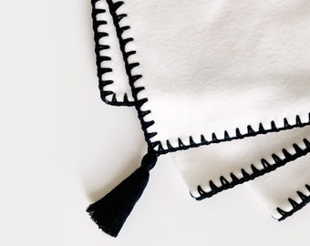 May include: White fleece blanket with black crocheted trim and a black tassel.