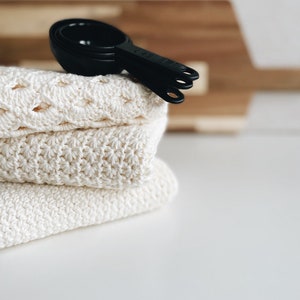 May include: A stack of three white crocheted dishcloths with black measuring cups on top.