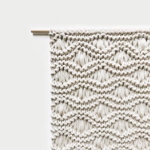 May include: A beige macrame wall hanging with a woven chevron pattern. The hanging is attached to a wooden dowel rod.