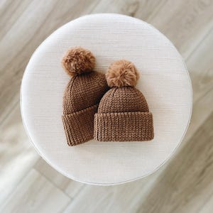 Peut inclure: Deux bonnets en tricot marron avec des pompons moelleux sur le dessus. Les bonnets sont posés sur une surface blanche.