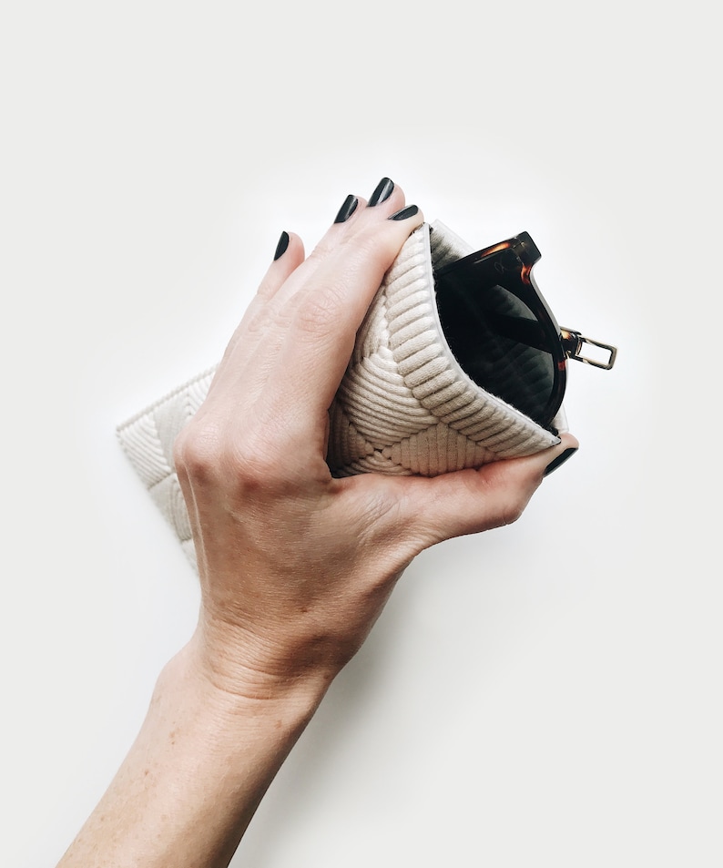May include: A hand holding a white quilted glasses case with a pair of brown and black sunglasses inside.