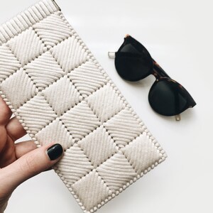 May include: A white crocheted eyeglass case with a diamond pattern. The case is held in a hand with black nail polish. A pair of black sunglasses with tortoise shell frames are lying on a white surface.