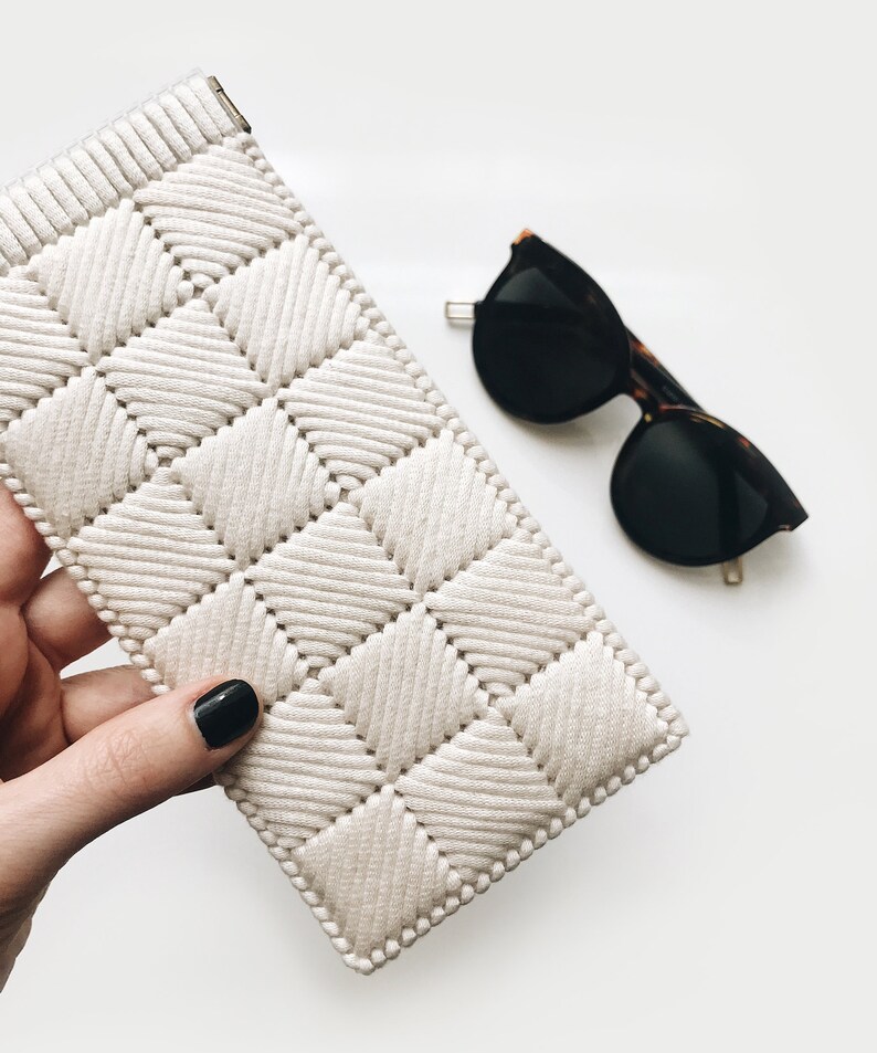 May include: A white crocheted eyeglass case with a diamond pattern. The case is held in a hand with black nail polish. A pair of black sunglasses with tortoise shell frames are lying on a white surface.
