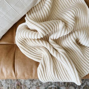 May include: Cream-colored, chunky knit throw blanket draped over a brown leather couch. The blanket has a ribbed texture and is folded to create visual interest. A cream-colored, square patterned pillow is visible in the upper left corner.