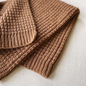 Könnte beinhalten: Eine braune gehäkelte Decke, die auf einer weißen Oberfläche gefaltet ist. Die Decke hat ein strukturiertes, geripptes Muster.