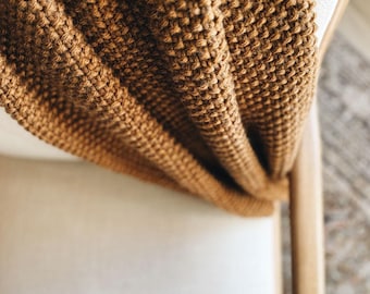 May include: A thick, textured knit blanket in a warm, earthy brown tone. The fabric features a dense, repeating stitch pattern that creates a raised, tactile surface. The blanket is draped over a light-colored seat, showing its heavy, folded drape.