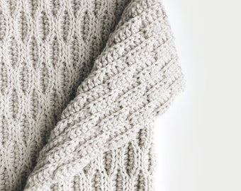 May include: A close-up of a beige crocheted blanket with a textured pattern. The blanket is folded in half and the edges are visible.
