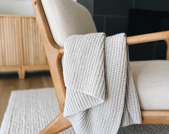 May include: A light beige knitted throw blanket draped over a mid-century modern style armchair. The chair has a light beige upholstered seat and back with a wooden frame. The blanket is a neutral color and has a ribbed texture. The chair is on a white rug.