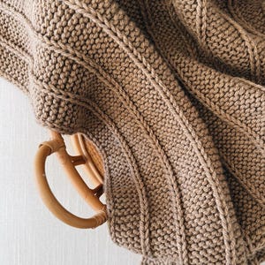 May include: A light brown knitted textile featuring a textured garter stitch pattern with vertical ribbed columns. The fabric is draped over a smooth, circular, light-colored wooden handle, creating folds and layers in the material.