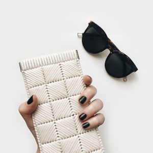 May include: A white woven clutch bag with a geometric pattern. The bag is held in a hand with black nail polish. A pair of black sunglasses with tortoise shell frames are in the background.