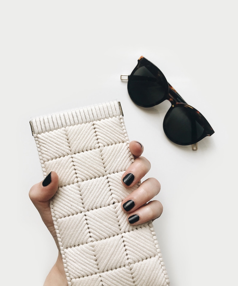 May include: A white woven clutch bag with a geometric pattern. The bag is held in a hand with black nail polish. A pair of black sunglasses with tortoise shell frames are in the background.