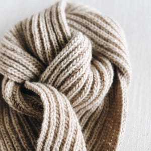 Pode incluir: Close-up de um cachecol bege de malha canelada. O cachecol está dobrado e amarrado, mostrando a textura do fio grosso. A cor neutra e o design simples sugerem um acessório aconchegante e versátil.