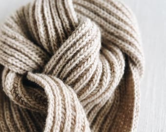 May include: Close-up of a beige, ribbed-knit scarf. The scarf is folded and knotted, showcasing the texture of the thick yarn. The neutral color and simple design suggest a cozy, versatile accessory suitable for various outfits.