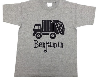 May include: A gray t-shirt featuring a black graphic of a garbage truck. The name "Benjamin" is printed in black script below the truck. The shirt has a crew neck and short sleeves.