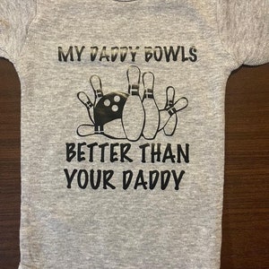 May include: Gray baby onesie with black text that reads "My Daddy Bowls Better Than Your Daddy" with a graphic of a bowling ball and pins.
