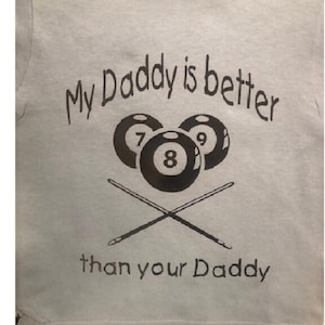 Puede incluir: Una camiseta azul claro con un gráfico negro de tres bolas de billar y tacos de billar cruzados. El texto "My Daddy is better than your Daddy" está impreso en la camiseta.