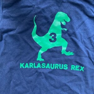 May include: A navy blue t-shirt with a green dinosaur graphic and the text "KARLASAURUS REX" and the number "3" on the dinosaur.
