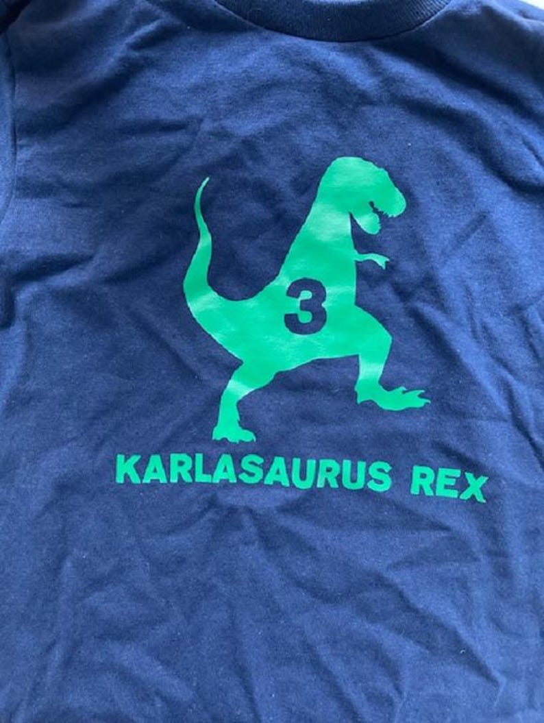 May include: A navy blue t-shirt with a green dinosaur graphic and the text "KARLASAURUS REX" and the number "3" on the dinosaur.