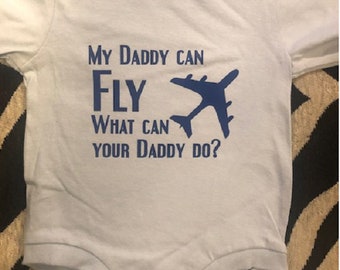 Peut inclure: Une grenouillère bébé bleu clair avec le texte "My Daddy Can Fly What Can Your Daddy Do?" en bleu foncé. Un graphique d'avion bleu est à côté du texte.