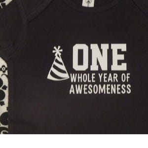 Pode incluir: Body de beb&eacute; preto com texto e gr&aacute;ficos brancos. O texto diz "ONE WHOLE YEAR OF AWESOMENESS" com um gr&aacute;fico de chap&eacute;u de festa. O body tem decote redondo e mangas curtas.