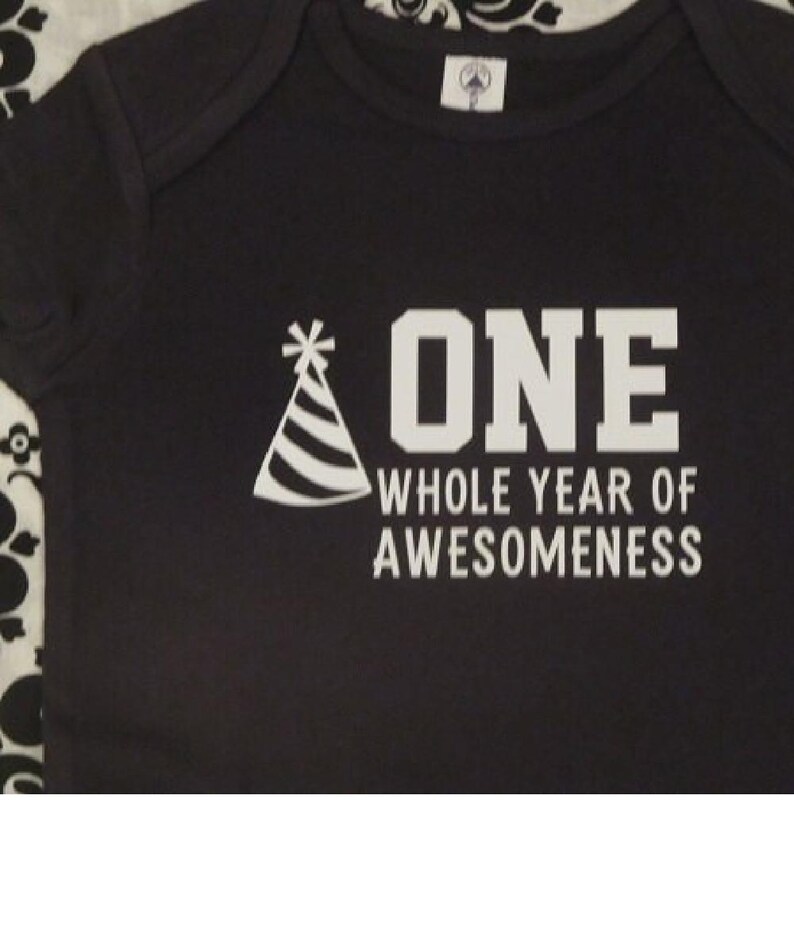 Pode incluir: Body de beb&eacute; preto com texto e gr&aacute;ficos brancos. O texto diz "ONE WHOLE YEAR OF AWESOMENESS" com um gr&aacute;fico de chap&eacute;u de festa. O body tem decote redondo e mangas curtas.