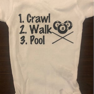 Puede incluir: Un body blanco para bebé con texto negro que dice "1. Crawl 2. Walk 3. Pool" y un gráfico de tres bolas de billar y dos tacos de billar cruzados.