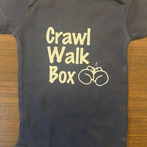 May include: A dark blue baby onesie with the text "Crawl Walk Box" and a pair of boxing gloves printed in white.