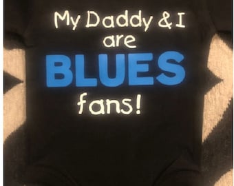 Può includere: Body nero con testo bianco che dice "My Daddy & I are BLUES fans!" La parola "BLUES" è in blu.