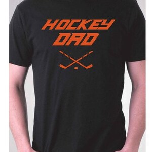 K&ouml;nnte beinhalten: Schwarzes T-Shirt mit dem Text "HOCKEY DAD" in Orange, darunter zwei gekreuzte Hockeyschl&auml;ger.