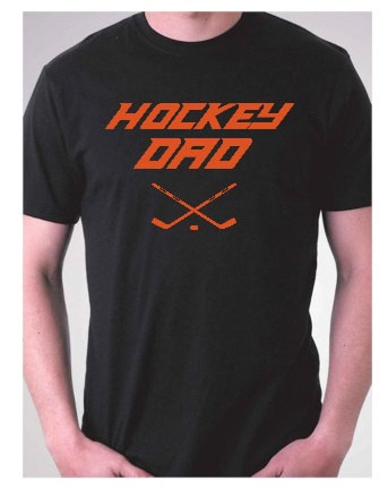 K&ouml;nnte beinhalten: Schwarzes T-Shirt mit dem Text "HOCKEY DAD" in Orange, darunter zwei gekreuzte Hockeyschl&auml;ger.