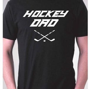 K&ouml;nnte beinhalten: Schwarzes T-Shirt mit wei&szlig;em Text "Hockey Dad" und einer Grafik von zwei gekreuzten Hockeyschl&auml;gern.