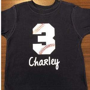 May include: A dark blue t-shirt with a white baseball-shaped number 3 and the name "Charley" written in white below it.