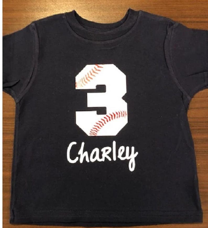May include: A dark blue t-shirt with a white baseball-shaped number 3 and the name "Charley" written in white below it.