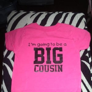 May include: Pink t-shirt with black text that reads "I'm going to be a BIG COUSIN".