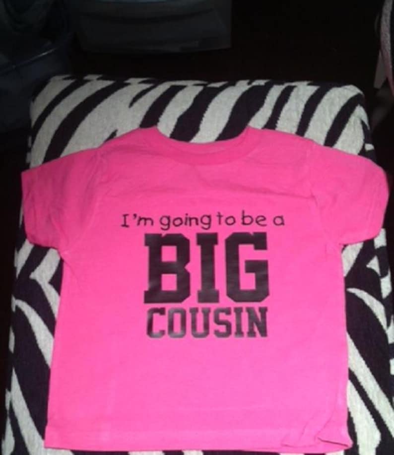 May include: Pink t-shirt with black text that reads "I'm going to be a BIG COUSIN".