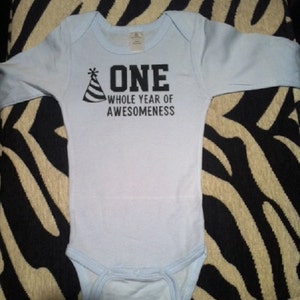 Pode incluir: Um body de beb&ecirc; azul claro com mangas compridas. O texto "ONE WHOLE YEAR OF AWESOMENESS" est&aacute; impresso em preto, acompanhado de um gr&aacute;fico de chap&eacute;u de festa. Este body &eacute; projetado para o primeiro anivers&aacute;rio de um beb&ecirc;.